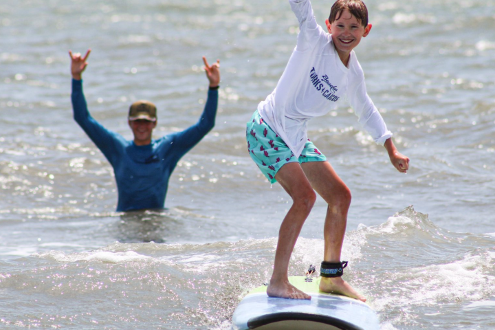 Child surfing on small wave while smiling adult raises hands in background.