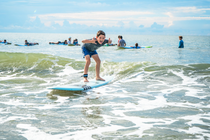Child surfing a small wave with others in the background on a sunny day.