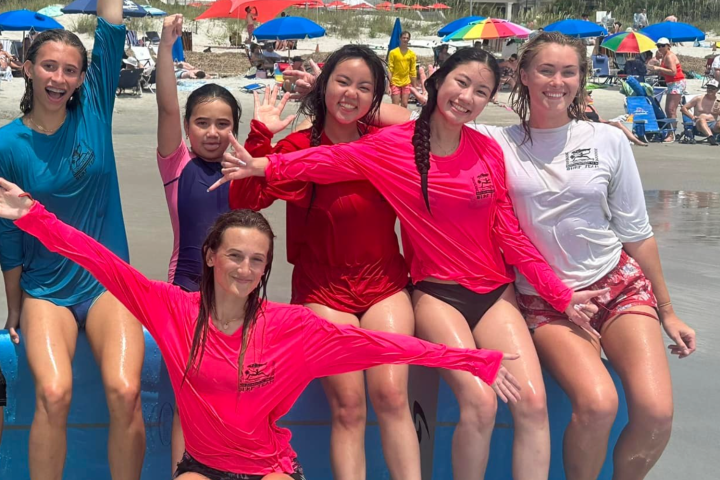 Six people in colorful shirts pose happily on a beach with umbrellas and buildings in the background.