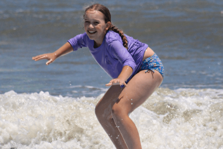 Girl in purple top surfing a wave on a sunny day.