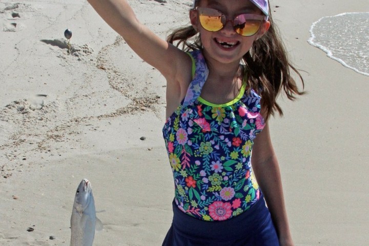 Girl holding a fish on a line at the beach, wearing colorful swimsuit and sunglasses.