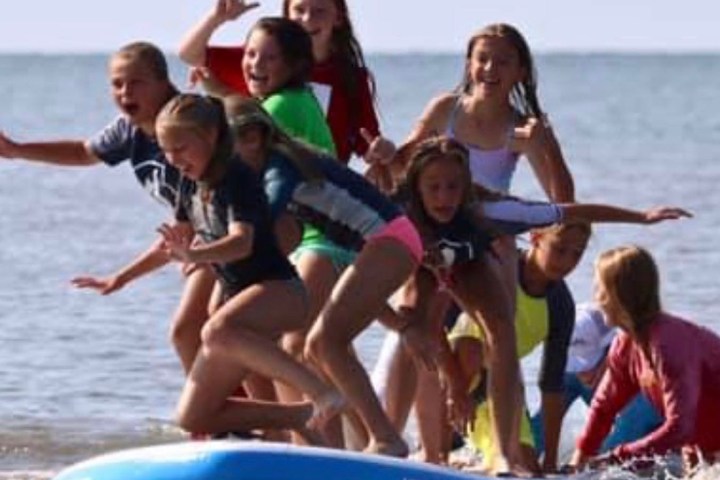 Group of kids balancing on surfboard in shallow ocean water, enjoying sunny day.