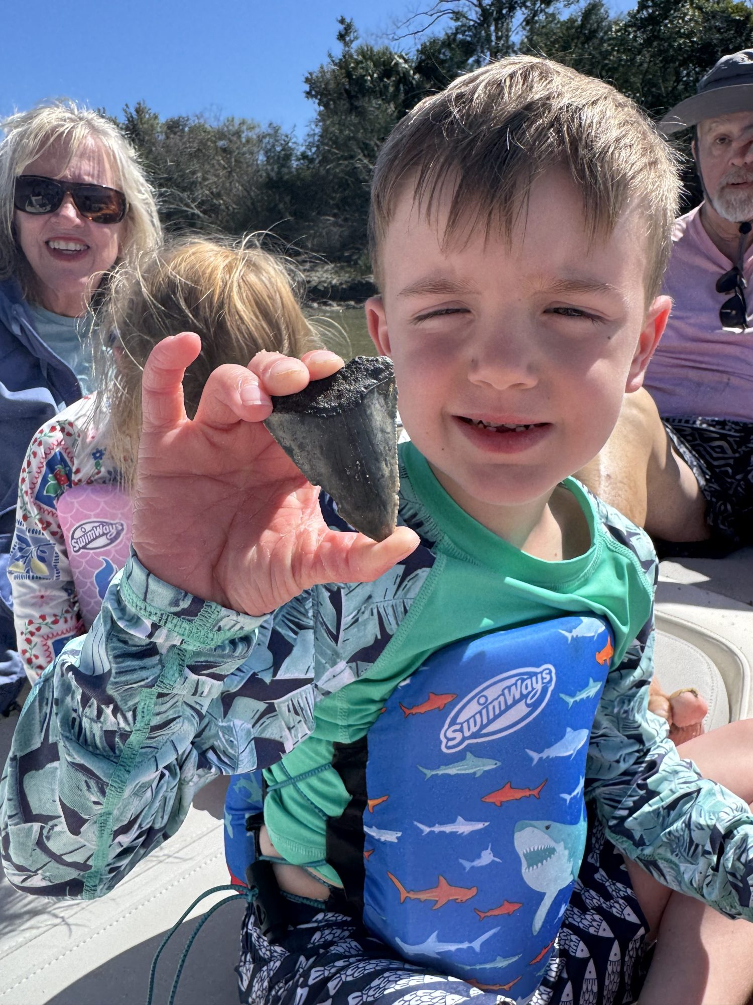Child in swim gear holding a large fossilized tooth, surrounded by family in a sunny outdoor setting.